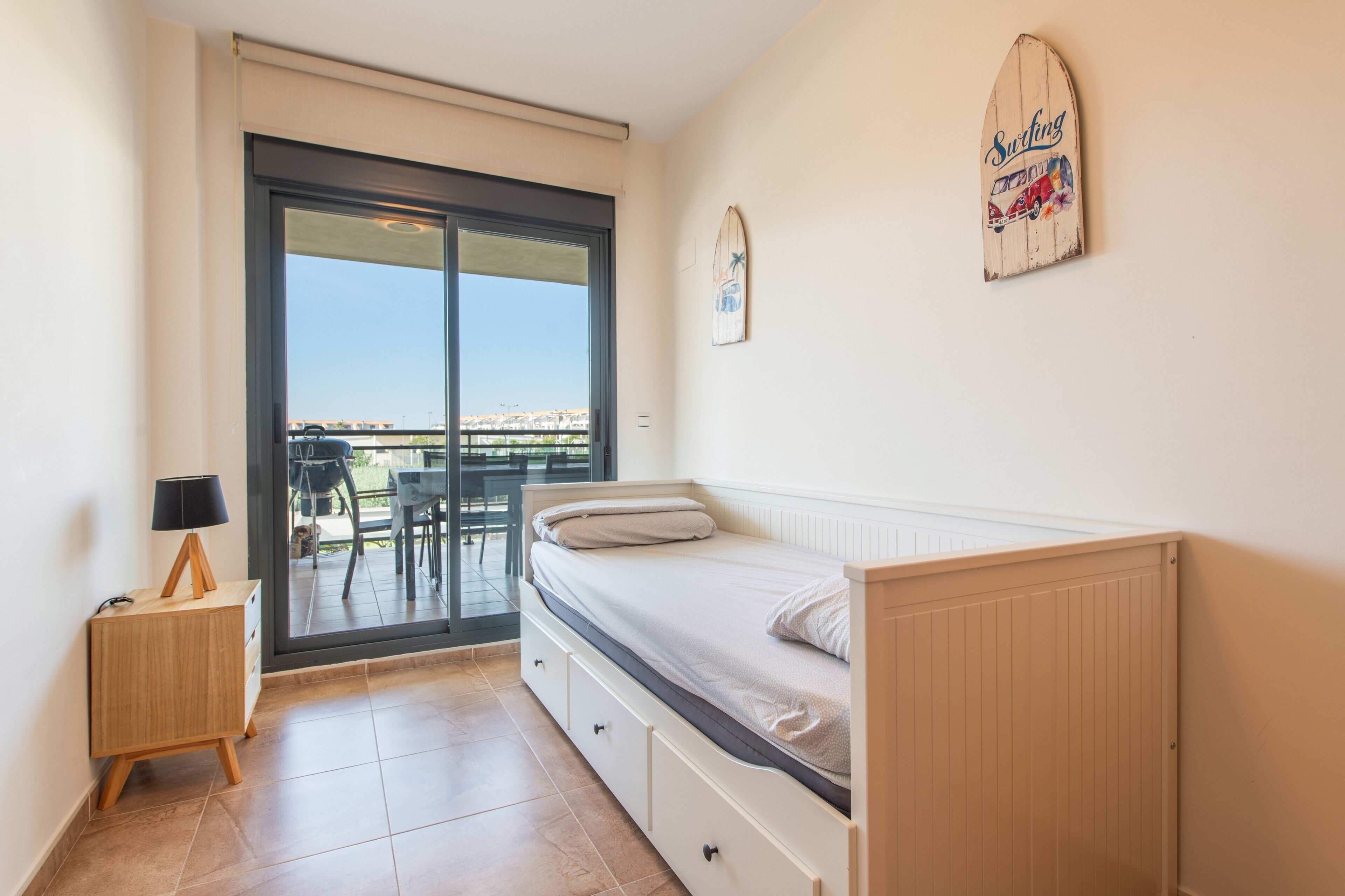 Un dormitorio con una cama, una mesita de noche, una lámpara, un ventanal corredero a un balcón y una decoración de tabla de surf en la pared.