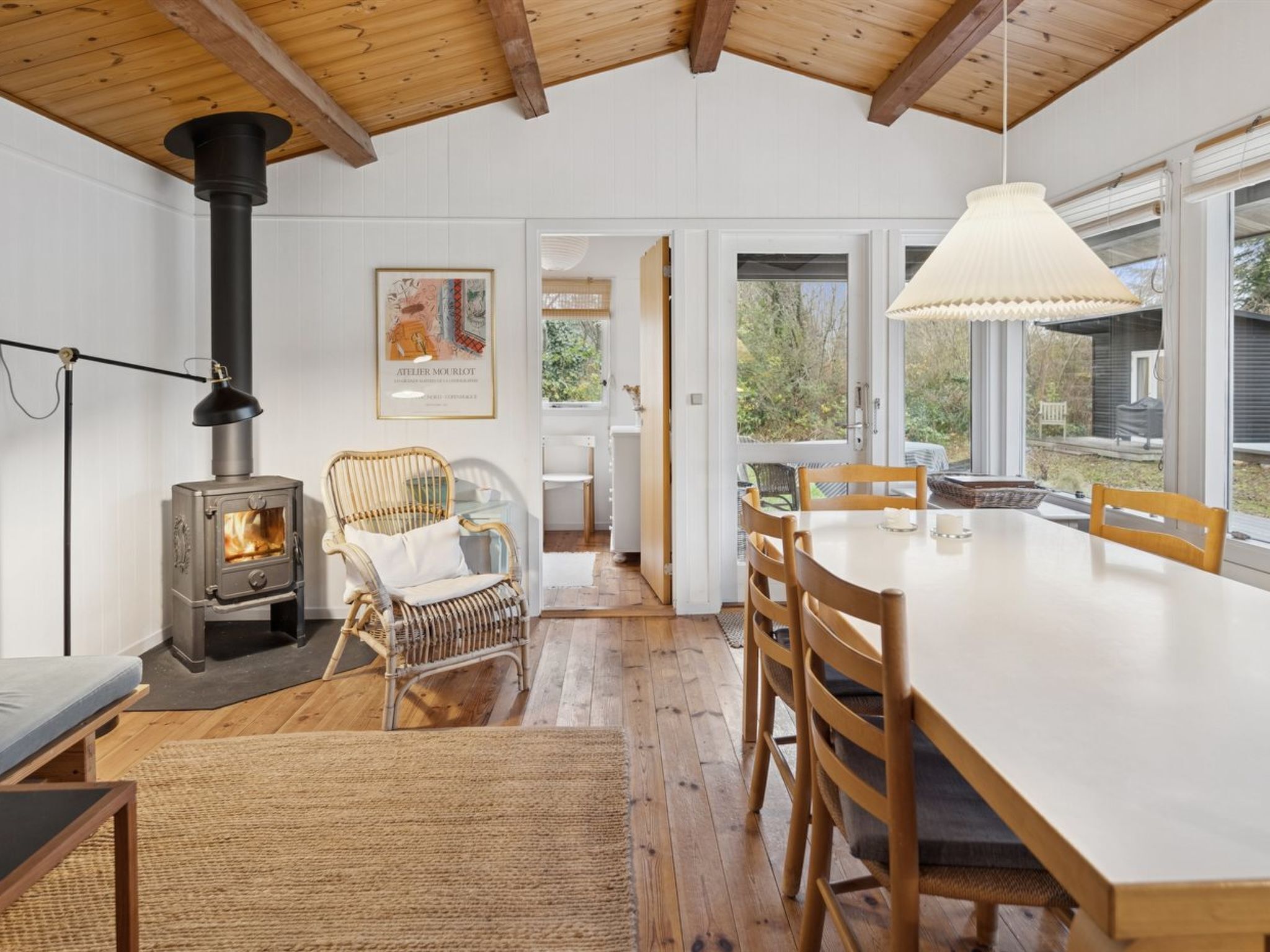 A cozy interior with a wood-burning stove, wicker chairs, a dining table, and large windows.