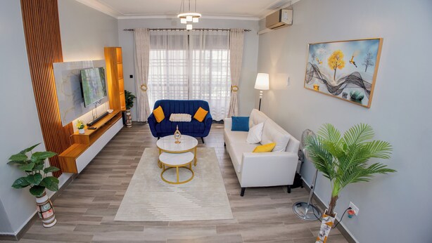 A modern living room with a sofa, a blue armchair, a coffee table, and a TV on a wooden console. - 2-bedroom apartment in peaceful Kampala with AC for relaxing stays (Kampala)