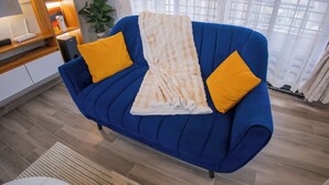 A blue sofa with a white throw blanket and two yellow pillows in a room with wooden flooring and a window with sheer curtains. - 2-bedroom apartment in peaceful Kampala with AC for relaxing stays (Kampala)
