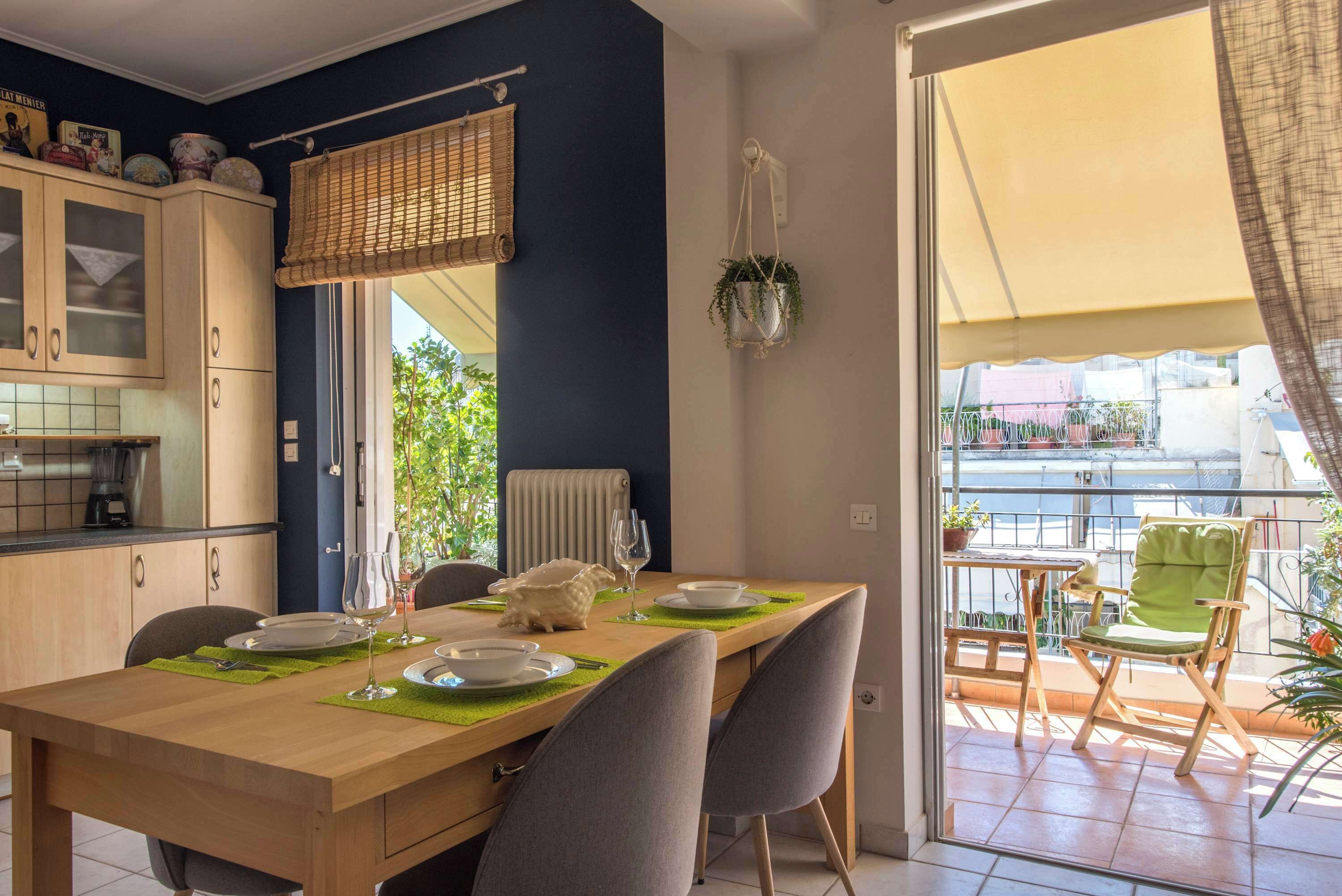 A dining area with a wooden table set for four, a kitchen with cabinets, and a balcony with a chair and plants.
