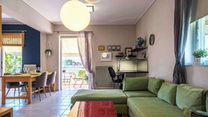 A living room with a green sofa, a wooden dining table with chairs, a red coffee table, and a large window with curtains. - Cozy Apartment with Beautiful Balcony (Athina)