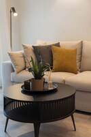 A round black table with a potted plant and a glass vase, a beige sofa with cushions, and a floor lamp in a living room.