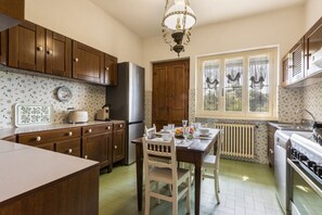 A kitchen with wooden cabinets, a dining table with chairs, a refrigerator, a stove, and a window with decorative curtains. - Villa la Leccia by MMega (Massarosa)
