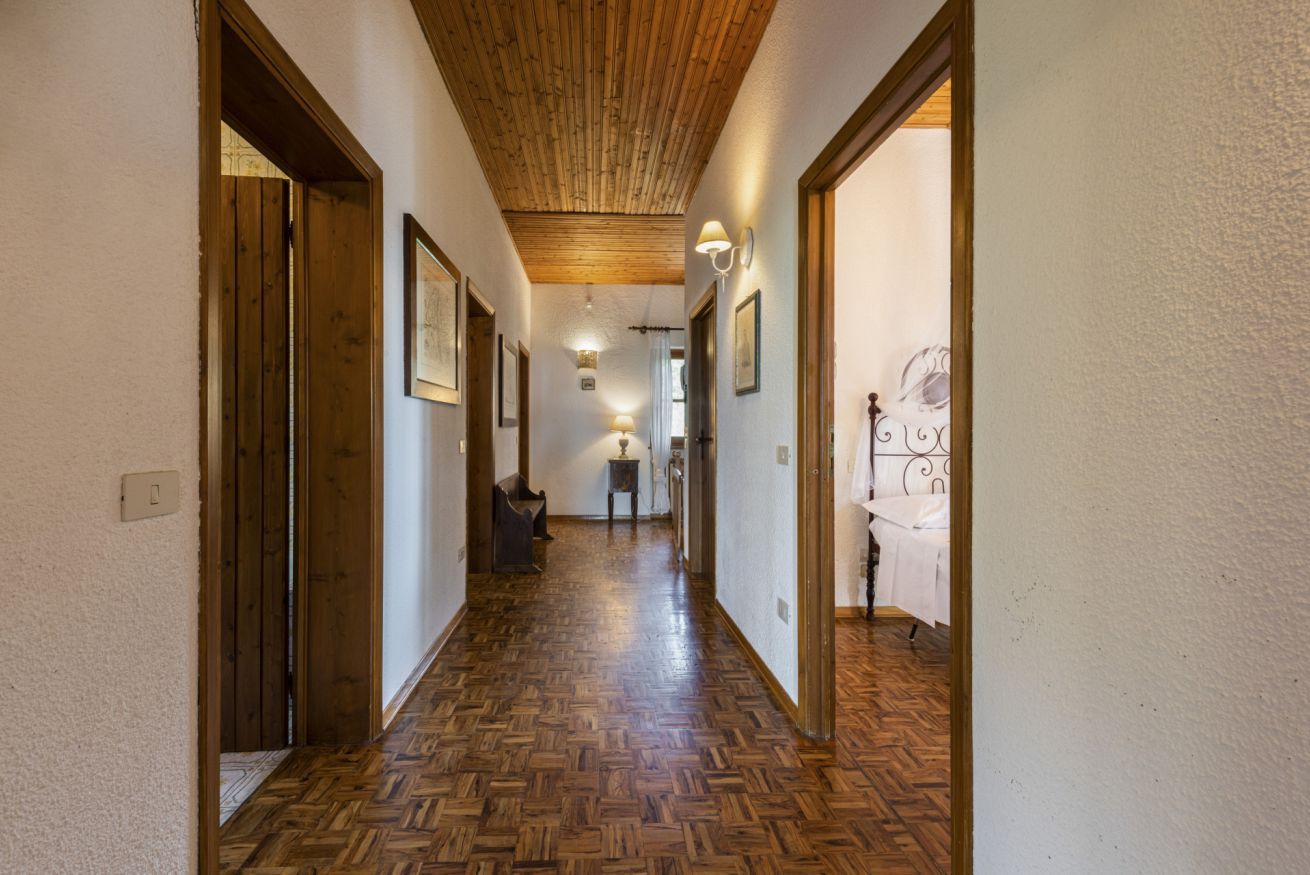 A hallway with wooden flooring, framed pictures on the walls, and a bed with a white bedspread in the background.