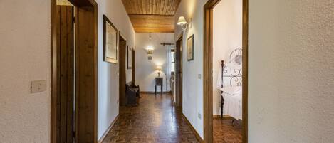 A hallway with wooden flooring, framed pictures on the walls, and a bed with a white bedspread in the background.