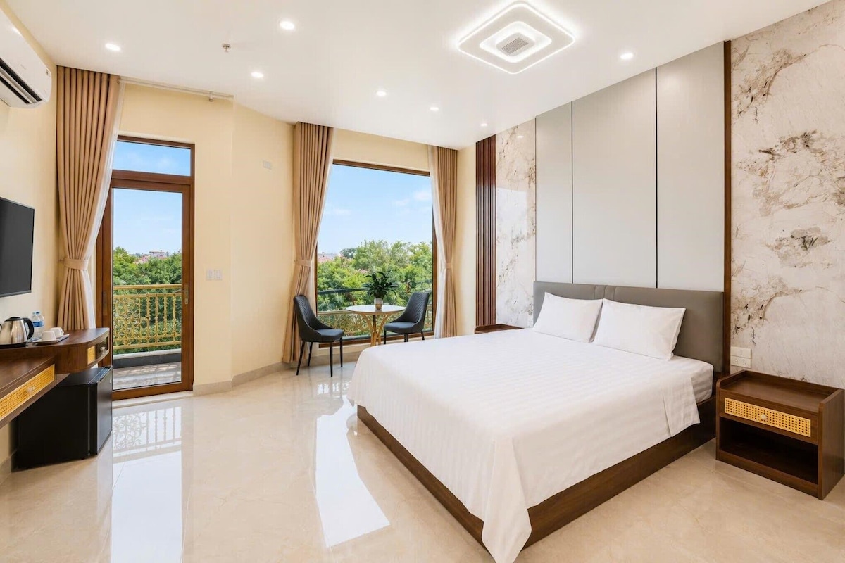 Habitación de hotel con cama, escritorio con televisor, balcón con sillas y vistas a zonas verdes.