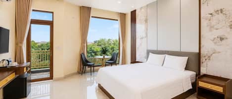 A hotel room with a bed, a desk with a TV, a balcony with chairs, and a view of greenery.