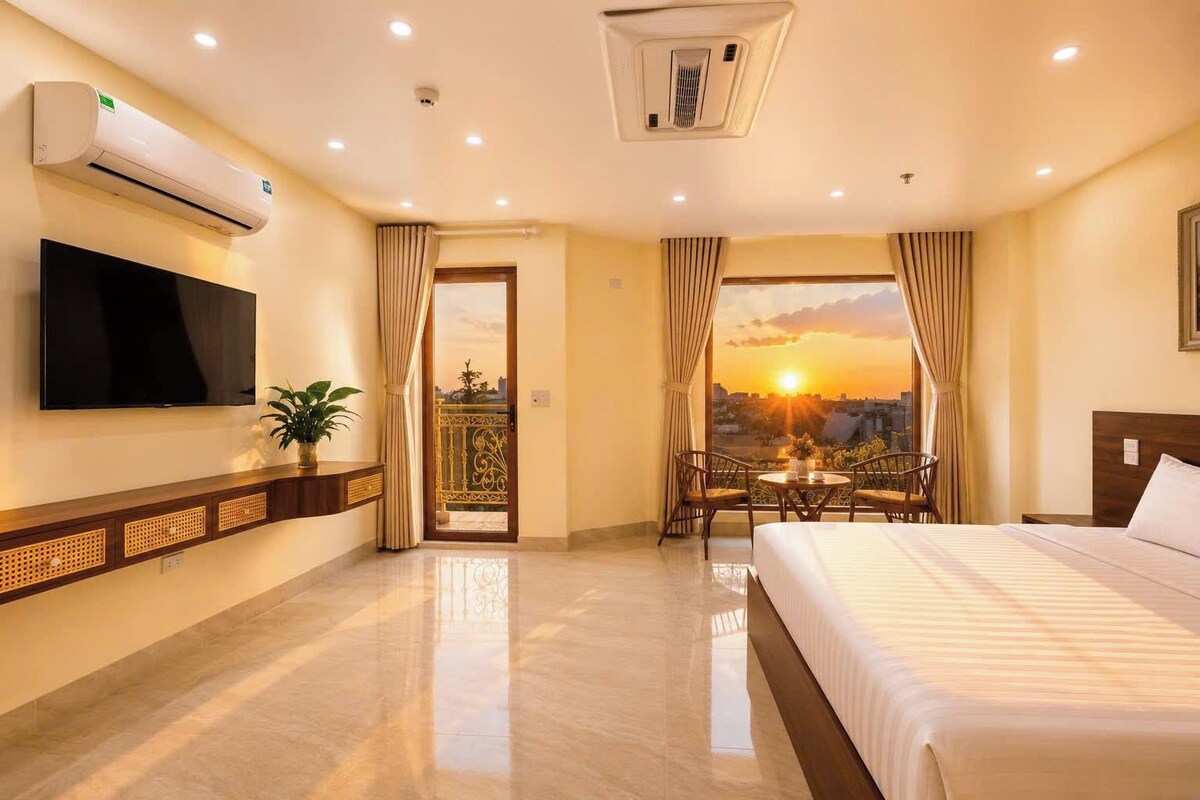 A hotel room with a bed, a TV, a balcony with a view of the sunset, and a small table with chairs.