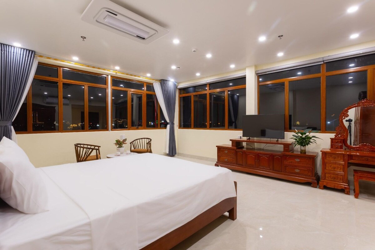 Una habitación de hotel con una cama grande, muebles de madera, un televisor de pantalla plana y ventanas amplias.