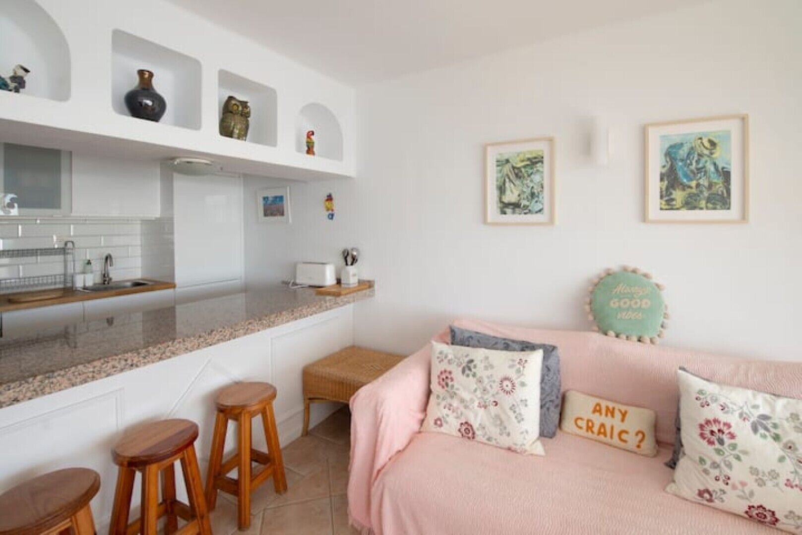 A kitchen with white cabinets, a granite countertop, and a sink. There are two wooden stools, a pink sofa with floral cushions, and a small table.