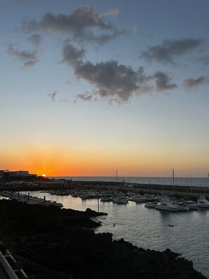 A marina with boats docked, a sunset in the background, and a rocky shoreline. - Roca Negra (Tías)