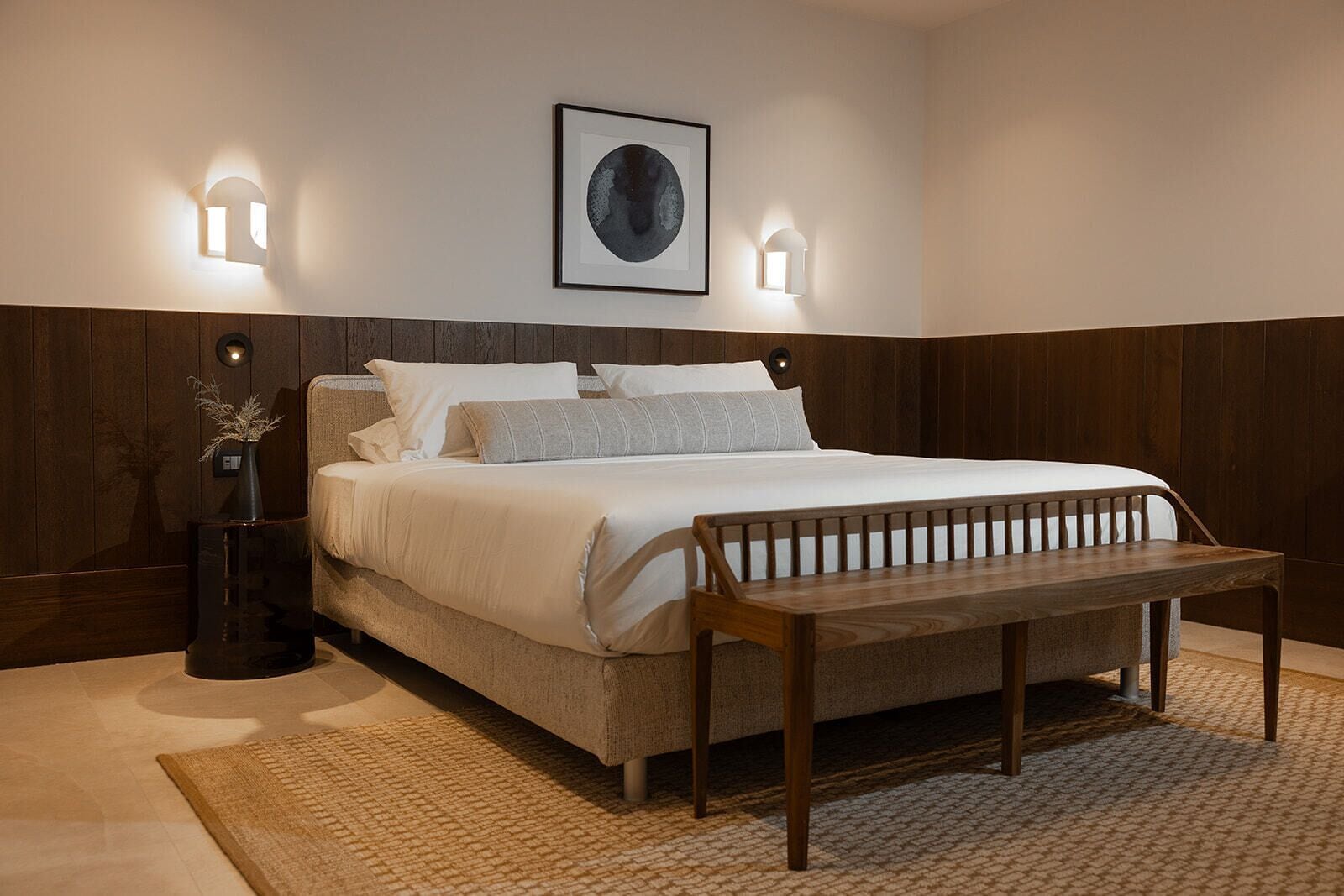 A bedroom with a bed, bedside lamps, a bench, a framed picture, and a vase with dried plants.
