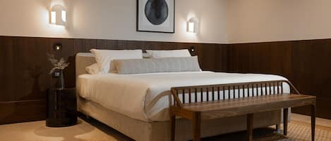 A bedroom with a bed, bedside lamps, a bench, a framed picture, and a vase with dried plants.