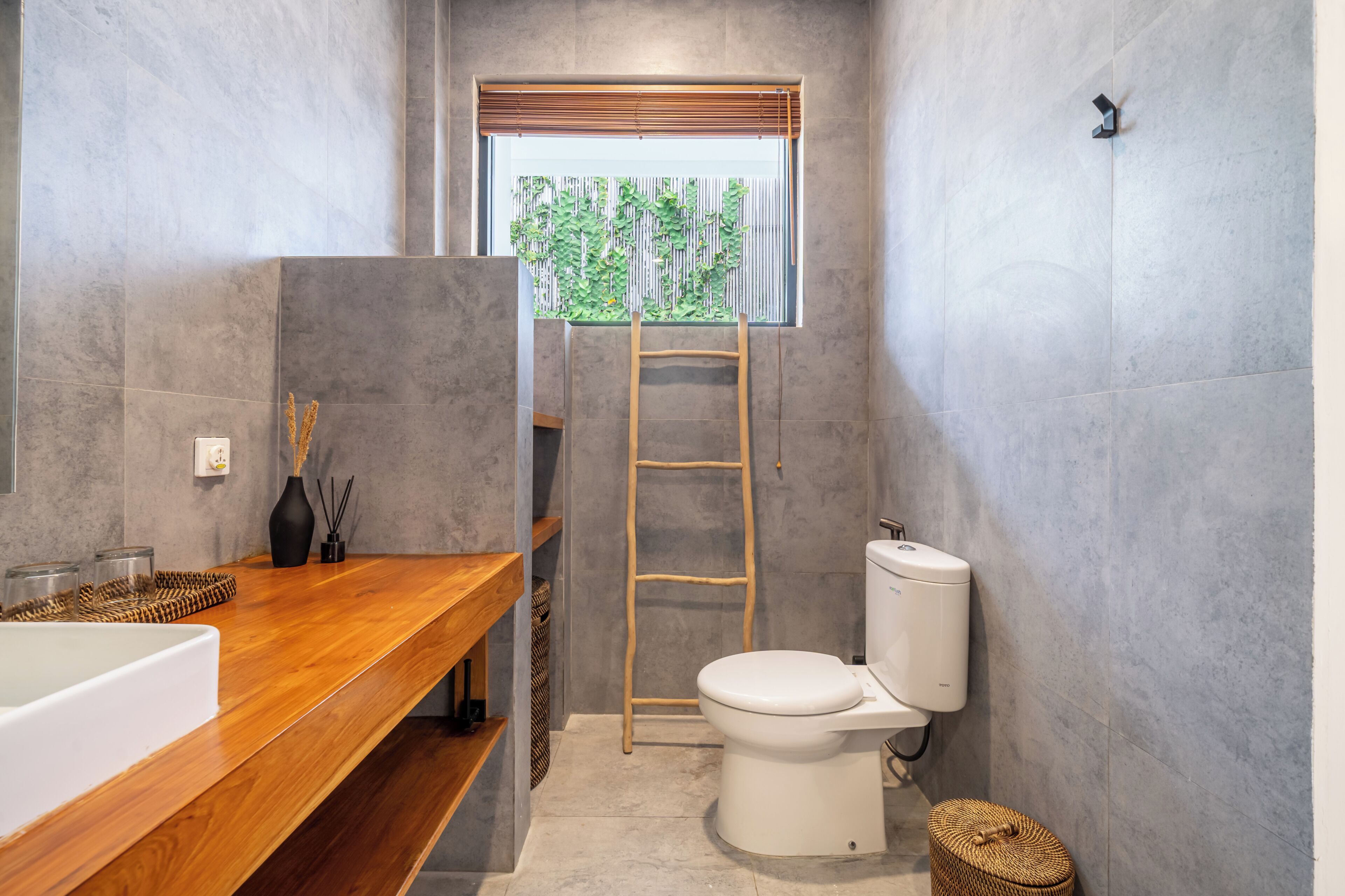 Ein Badezimmer mit Waschtisch aus Holz, einer weißen Toilette, einem Fenster mit Blick ins Grüne und einer Leiter.