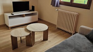 A living room with a sofa, two round wooden tables, a TV on a white stand, a radiator, and a window with a view of a building and a fence. - Little cocoon (Ferrières-en-Bray)