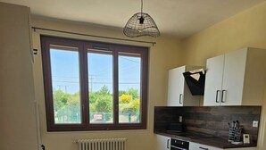 A kitchen with a dining table, chairs, a radiator, and a window with a view of greenery. - Little cocoon (Ferrières-en-Bray)