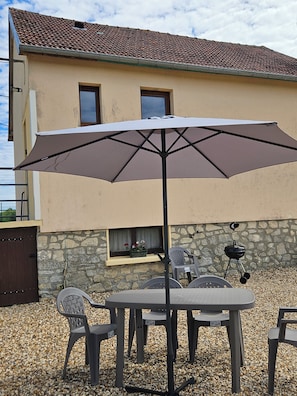 Une terrasse extérieure avec une table et des chaises, un grand parasol, un barbecue et un mur en pierre.