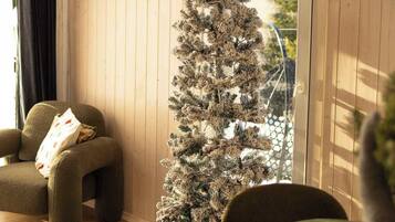A cozy room with a wooden ceiling, a framed sign on the wall, a Christmas tree, a sofa, a dining table with placemats, and a gift box.