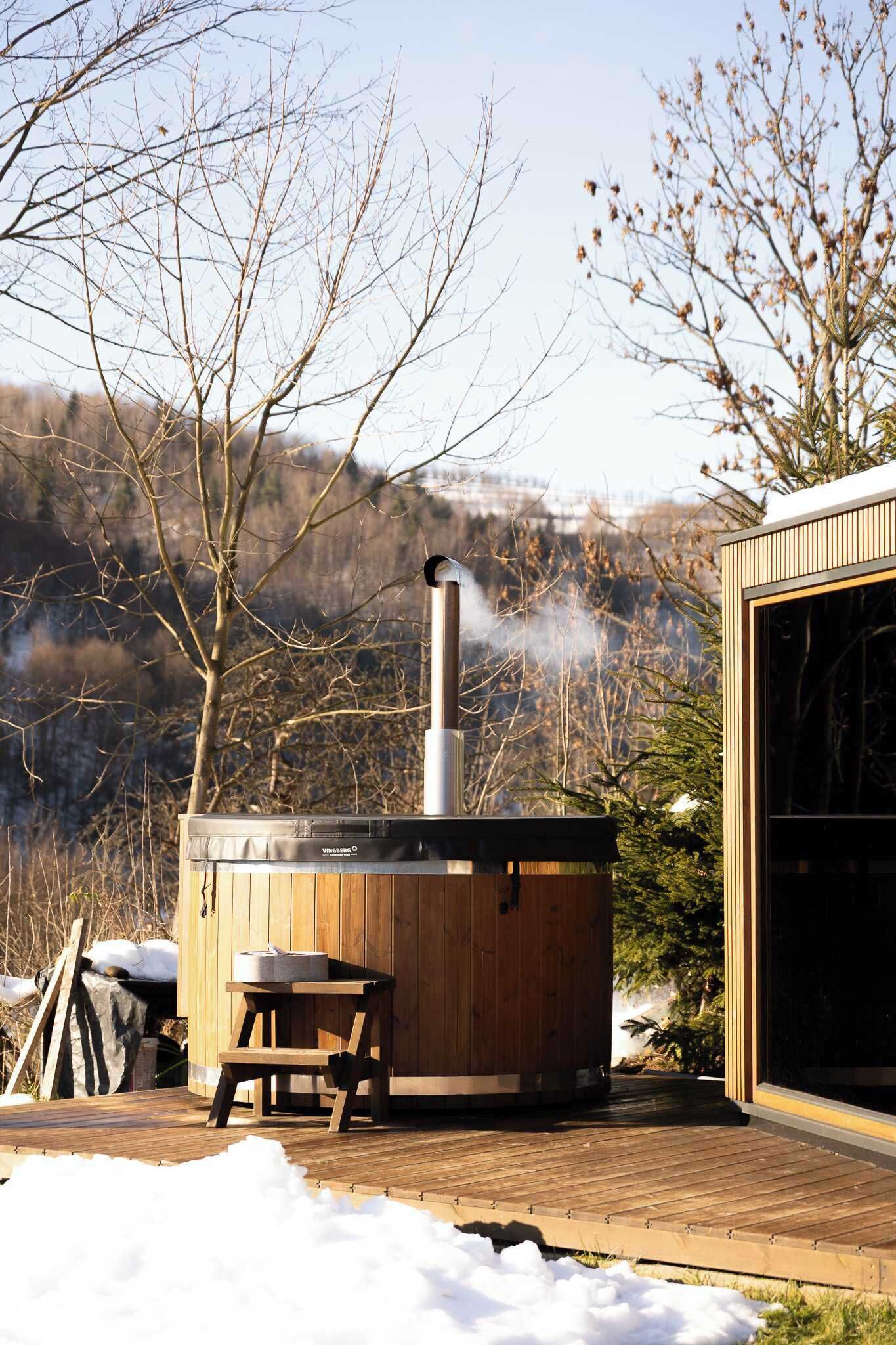A wooden hot tub with a chimney, a table, and a bench in a snowy outdoor setting.