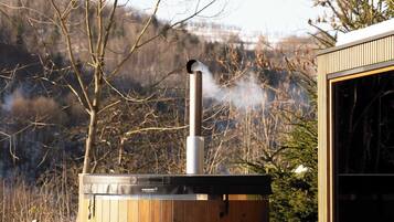A wooden hot tub with a chimney, a table, and a bench in a snowy outdoor setting.