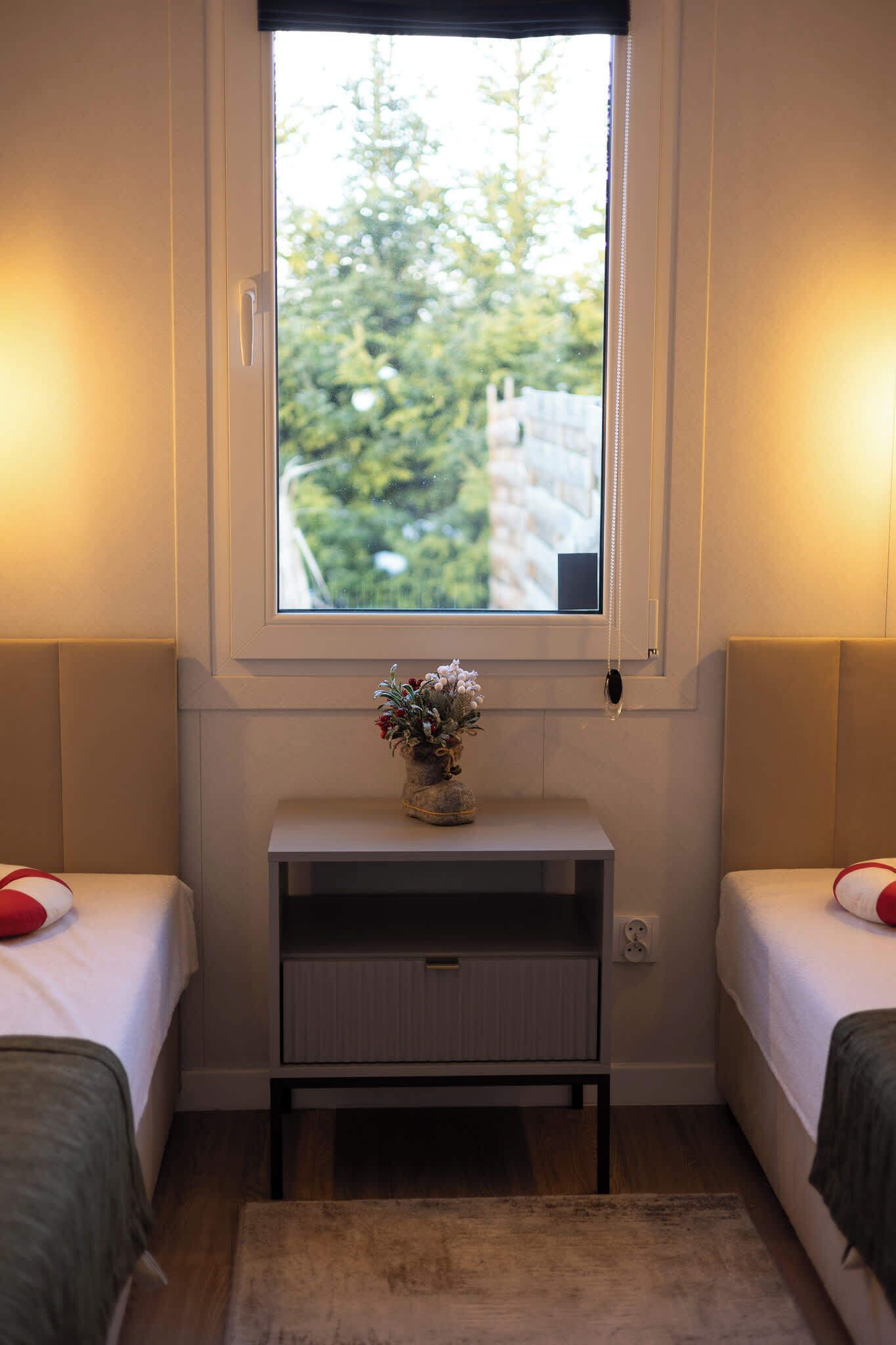 A bedroom with two beds, a bedside table with a vase, a window with a view of greenery, and a small rug.