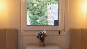 A bedroom with two beds, a bedside table with a vase, a window with a view of greenery, and a small rug.