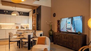 Uma sala de estar moderna com televisão, canto de jantar e cozinha ao fundo.