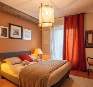 Hotellihuone, jossa on sänky, yöpöytälamppu, ikkuna verhoilla ja seinällä kehystettyjä kuvia.