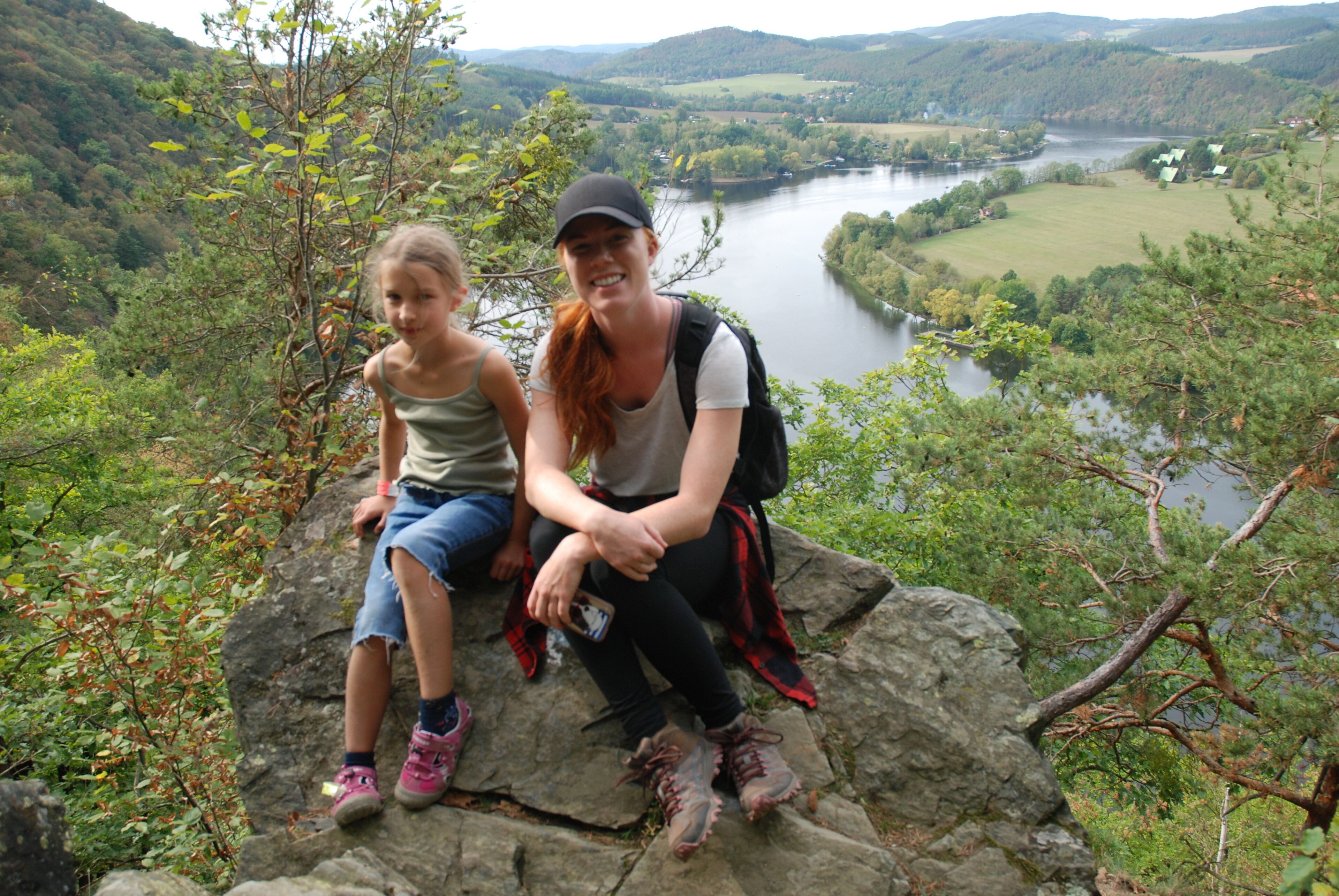 Zwei Personen sitzen auf einem Felsen, im Hintergrund fließt ein Fluss und erheben sich Hügel.