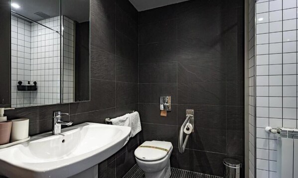 Un baño moderno con un lavamanos grande, inodoro y zona de ducha, con baldosas oscuras y un espejo.