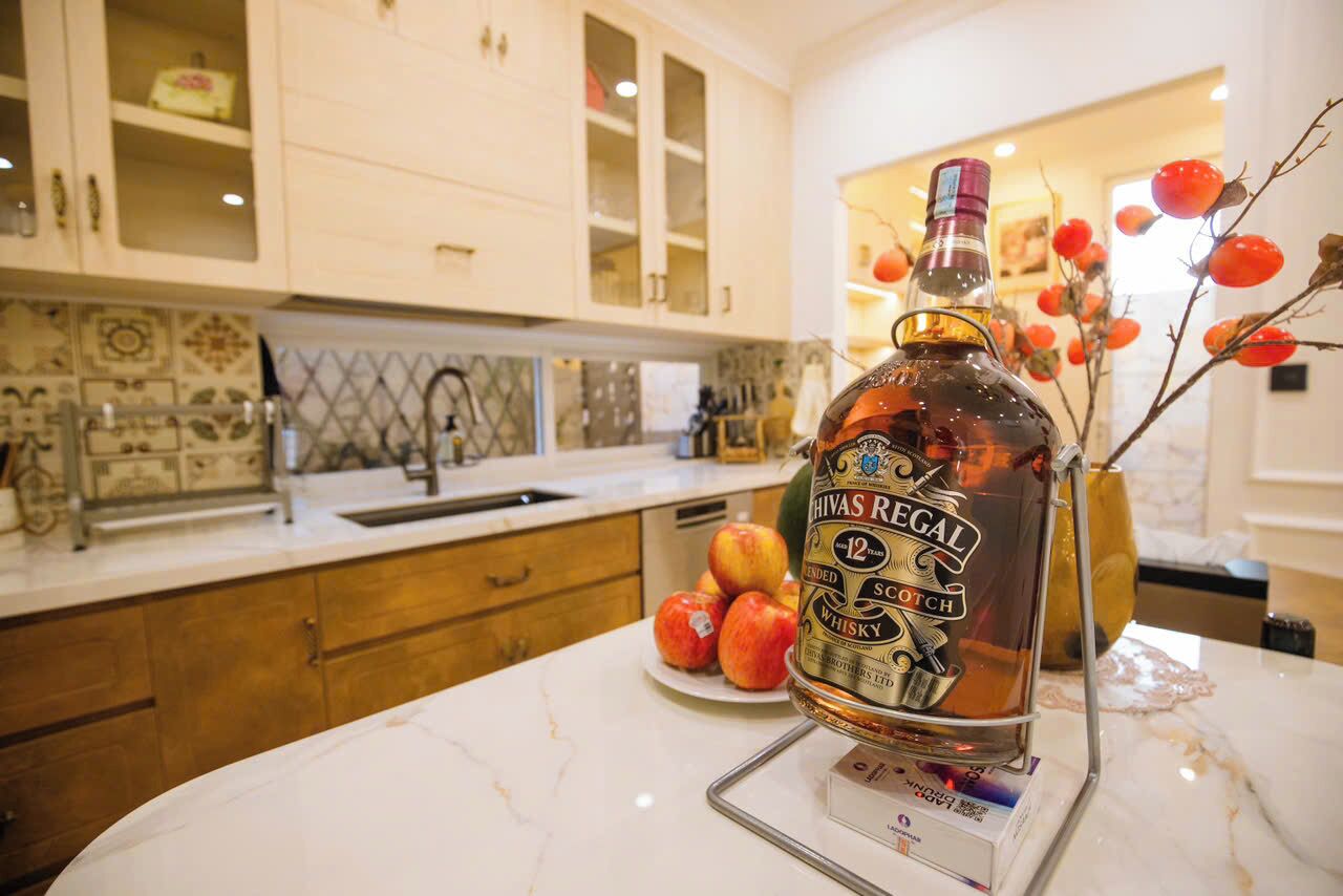 A bottle of Hivas Regal 12-year-old scotch whisky on a marble countertop in a kitchen with white cabinets and a tiled backsplash.