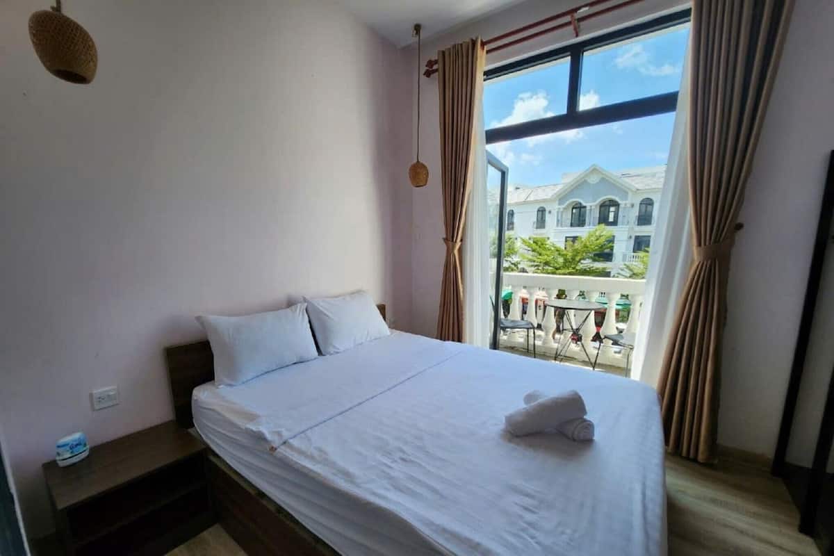 A hotel room with a bed, a small table, a window with curtains, and a view of buildings outside.