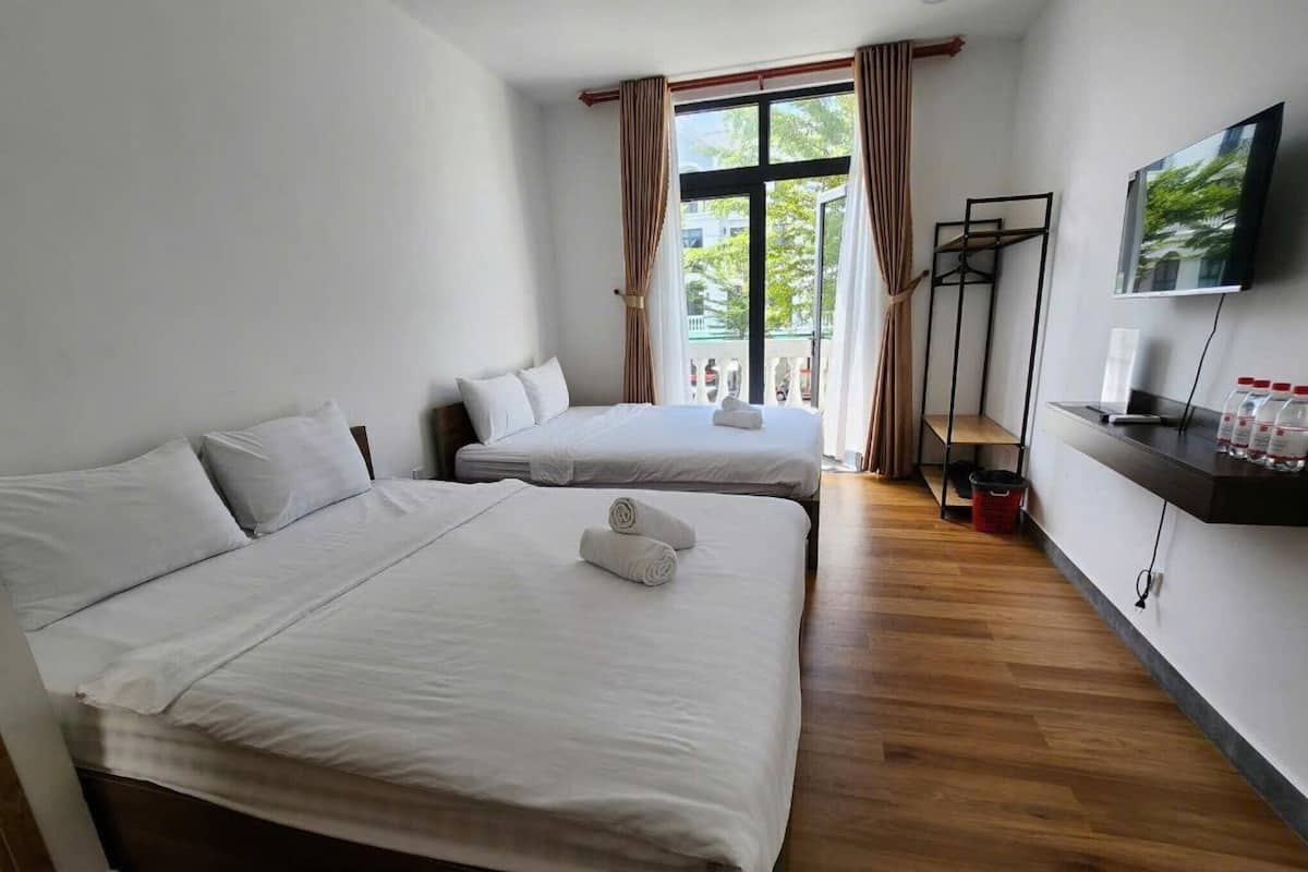 A hotel room with two beds, a TV, a shelf with bottles, and a window with curtains.