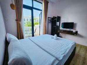 A hotel room with a bed, a TV, a small table with bottles, and a view of buildings and greenery outside.