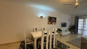 Un coin repas avec une table et des chaises blanches, un ventilateur de plafond, un téléviseur et une plante.