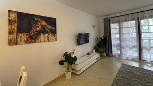 Un salon avec un tableau accroché au mur, un téléviseur à écran plat, une console blanche, une plante en pot et une grande fenêtre avec des rideaux.