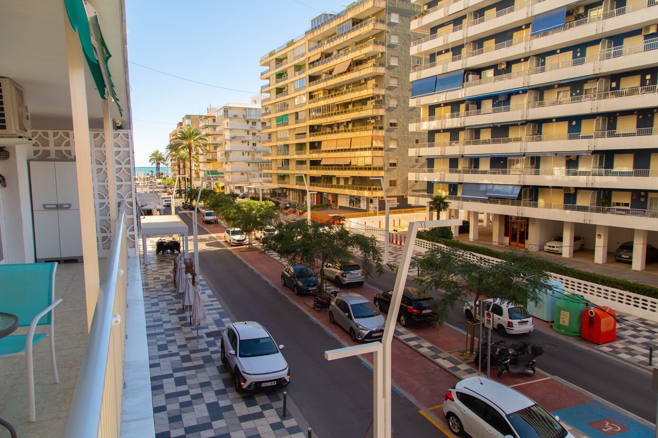 Um hotel de vários andares com varandas, calçada quadriculada, carros estacionados e palmeiras.