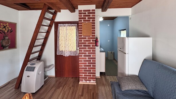 Une pièce avec un plafond en bois, une colonne en briques, un réfrigérateur blanc, un canapé bleu, une échelle en bois, des toilettes et une porte ornée de rideaux à motifs floraux.