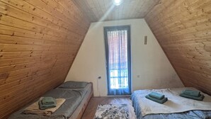 Une petite chambre mansardée, chaleureuse et accueillante, avec des murs en bois et un plafond en pente. Elle comprend deux lits simples avec des têtes de lit assorties, une petite fenêtre avec des rideaux et un tapis à motifs sur le plancher en bois.