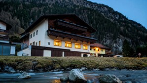 Et hotel i traditionel alpearkitektur, med en flod foran og et bjerglandskab i baggrunden.
