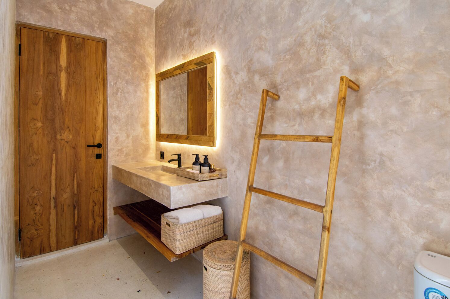 A bathroom with a wooden door, a marble sink, a mirror with a wooden frame, a wooden ladder, and wicker baskets.