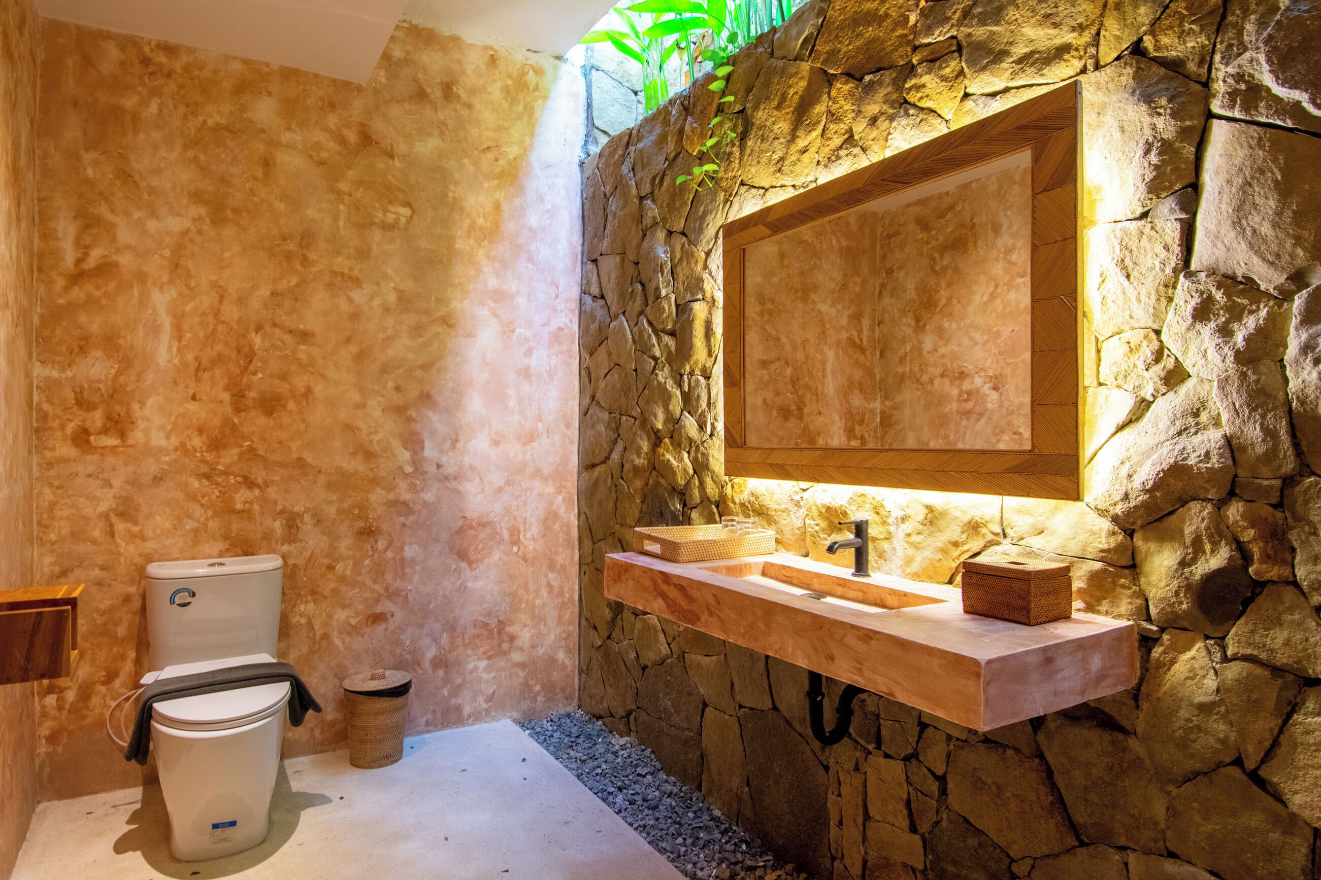 A bathroom with a stone wall, a wooden vanity, a toilet, and a small plant.