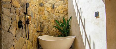 Un baño con paredes de piedra, una bañera independiente, cabezal de ducha y una planta en maceta.
