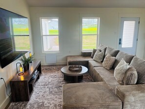 Ein Wohnzimmer mit einem großen Flachbildfernseher, einer Holz-Unterhaltungsecke, einem gemusterten Teppich, einem beigen Sofa mit Kissen und zwei Fenstern mit Blick auf eine grüne Landschaft.