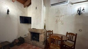 A room with a stone fireplace, a round wooden table with chairs, a mounted flat-screen TV, and an air conditioning unit.