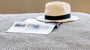 Un sombrero de paja sobre un tapete tejido, con una revista, con vistas a una playa con palmeras y el océano.