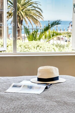 Ein Strohhut auf einer gewebten Matte mit einer Zeitschrift, mit Blick auf einen Strand mit Palmen und dem Meer.