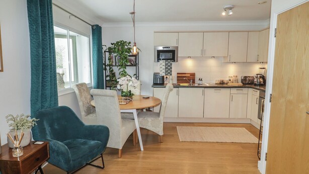 A modern kitchen with white cabinets, a dining area with a table and chairs, and a teal armchair. - Cornstore Cottage (Pembroke)