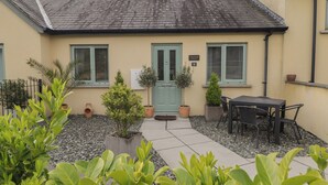 A building with a tiled roof, a door, and windows. There are potted plants and a table with chairs outside. - Cornstore Cottage (Pembroke)